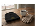 Black beanie with gold emblem, white mug with steaming coffee, and beige gloves on a wooden table.