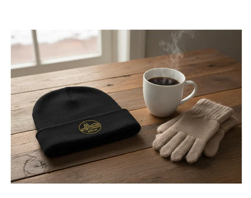 Black beanie with gold emblem, white mug with steaming coffee, and beige gloves on a wooden table.