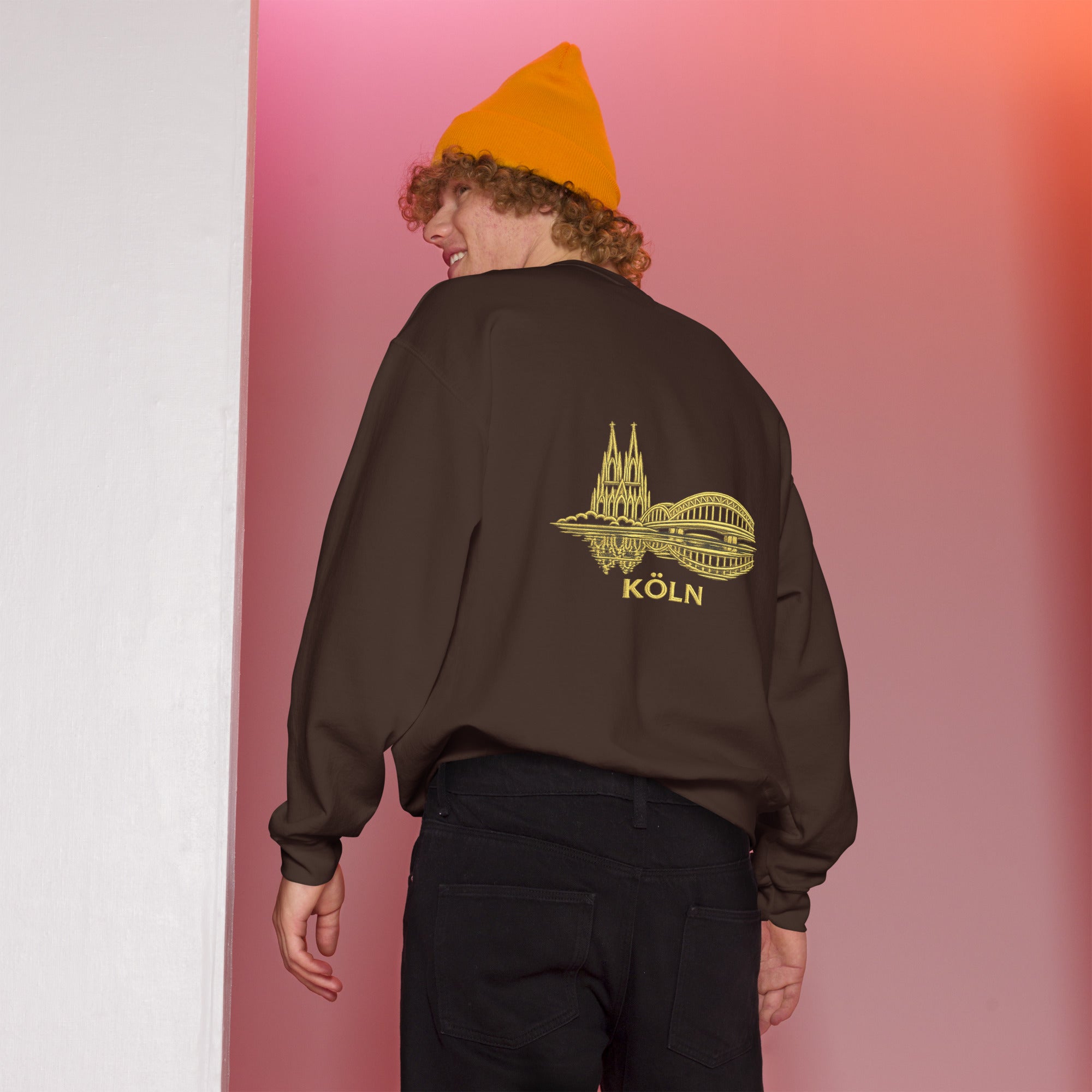 Person wearing a brown sweatshirt with a yellow 'Köln' design on the back, standing against a gradient pink and white background.