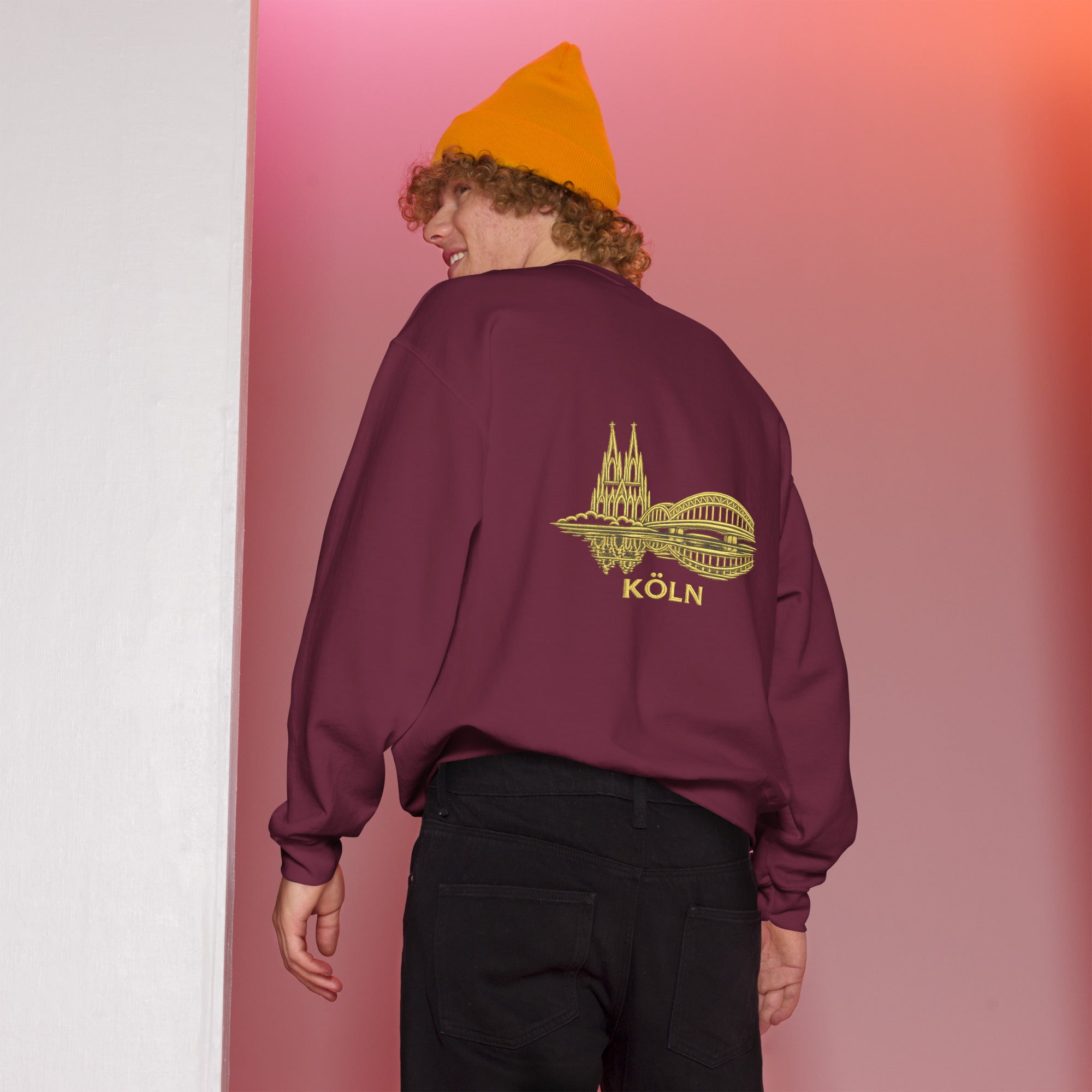 Person wearing a maroon sweatshirt with a 'Köln' design on the back, standing against a gradient pink and white background.